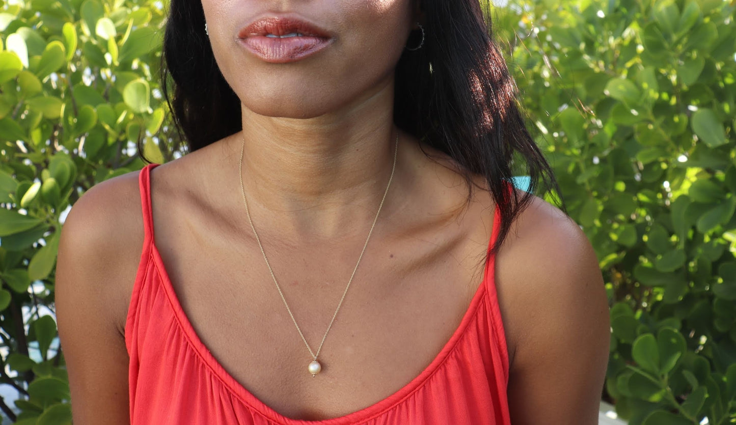 black woman with long brown hair and an orange shirt sitting in a garden and wearing the golden South sea pearl necklace from the wandering jewel