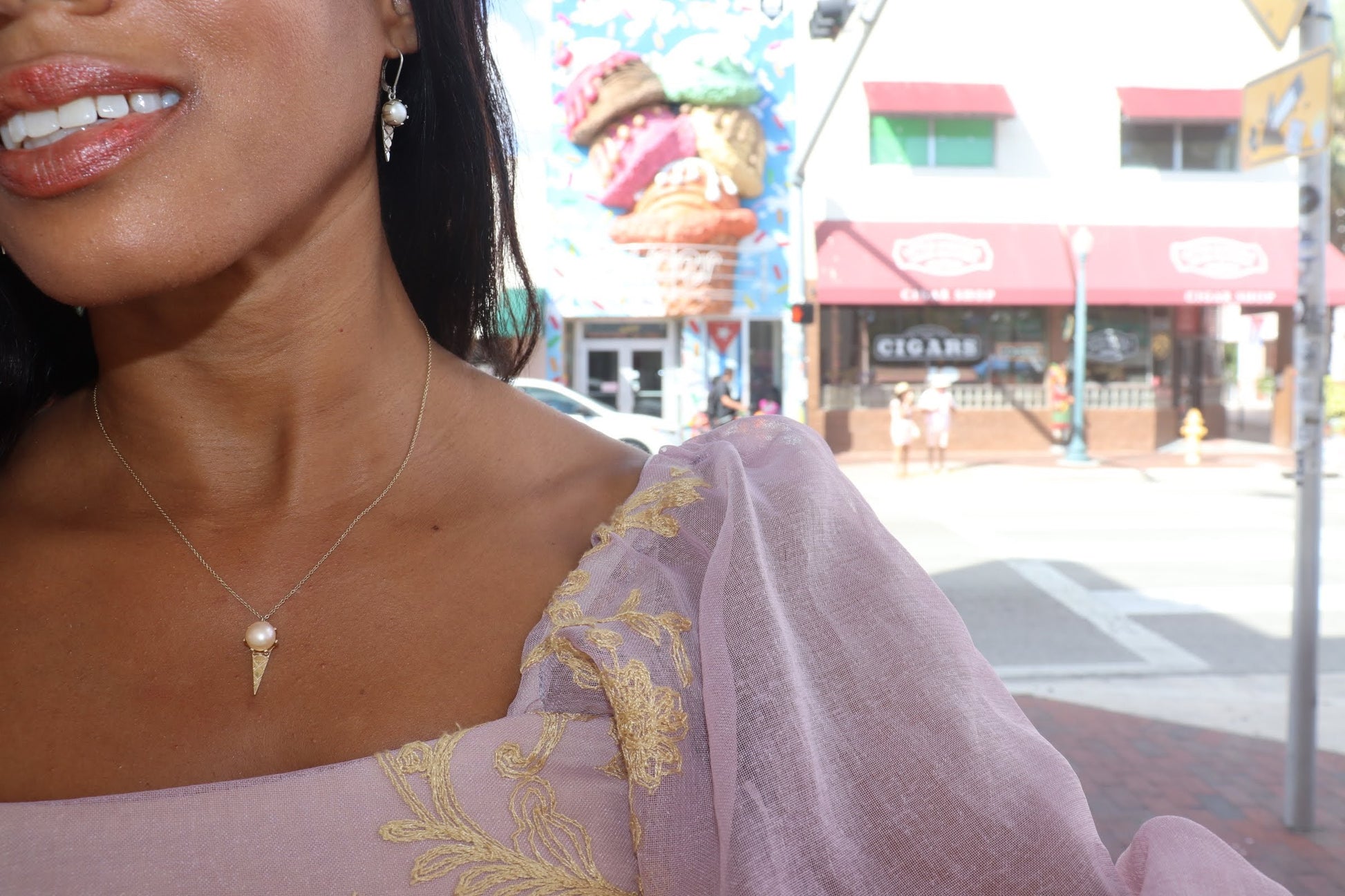 woman in a pink blouse standing in front of an ice cream shop and wearing the ice cream earrings and matching necklace from the wandering jewel