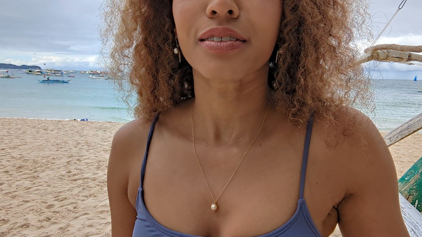 black woman with curly hair and purple bikini on beach wearing the golden South sea pearl pendant from the wandering jewel
