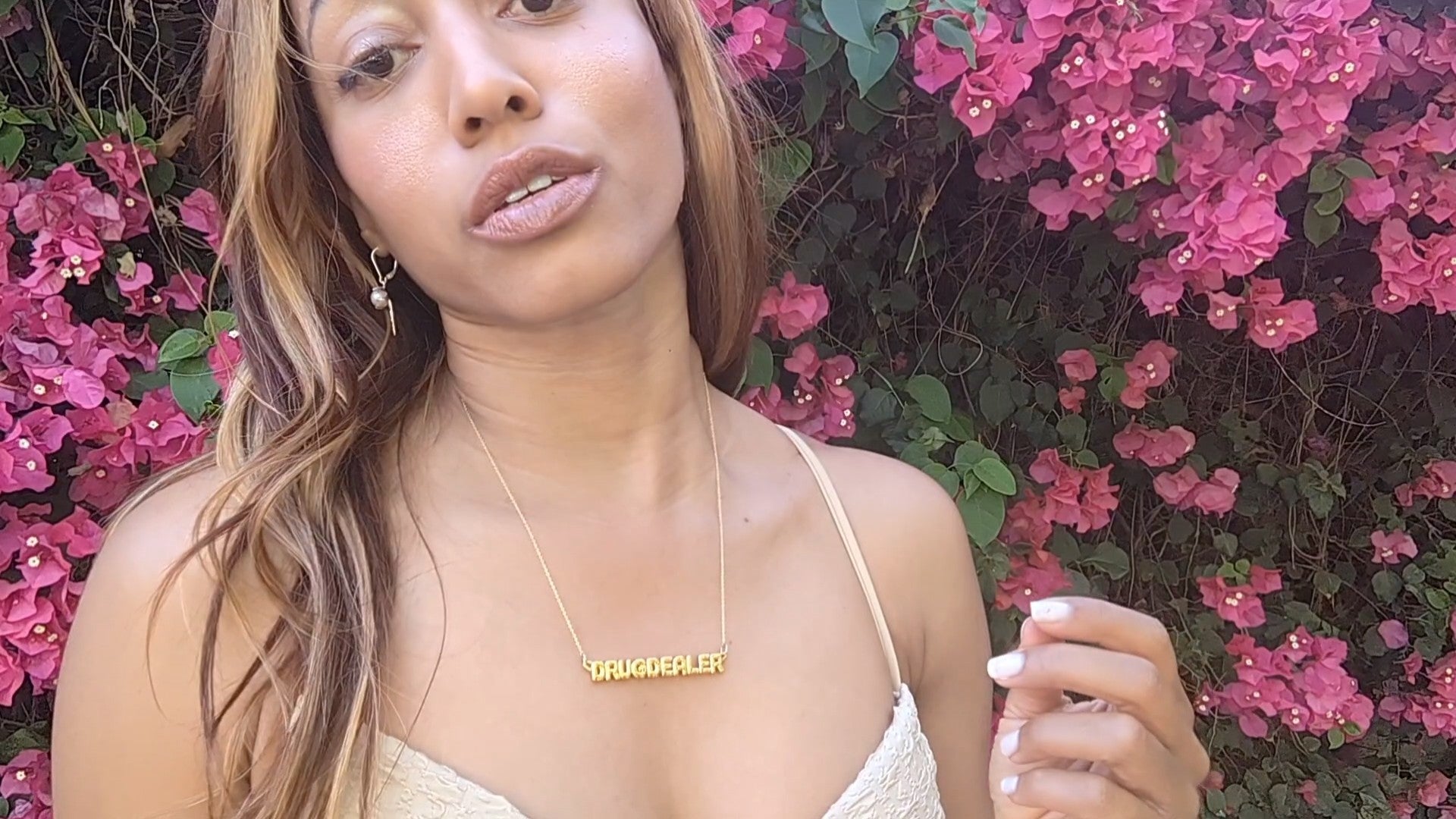 woman in tan bikini top standing in front of pink roses wearing the drug dealer nameplate necklace from the wandering jewel
