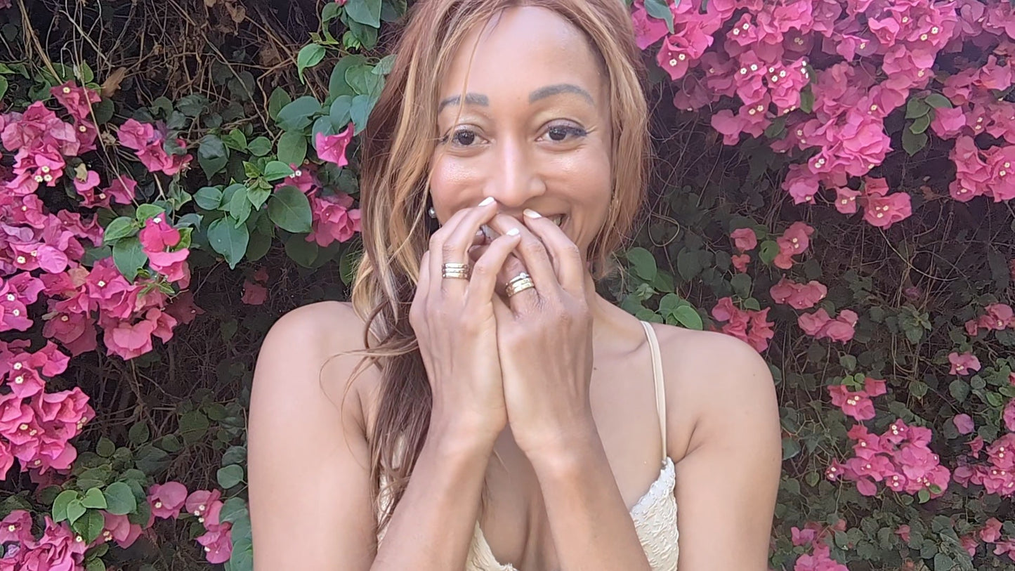 woman smiling in light brown tank top and standing in front of bright pink flowers wearing 6  18K solid gold disengagement rings with gray diamonds