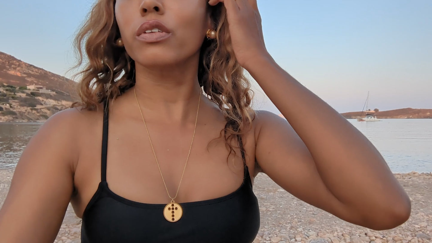 black woman in black bikini on Greek beach wearing the  Cross cutout coin from The Wandering Jewel