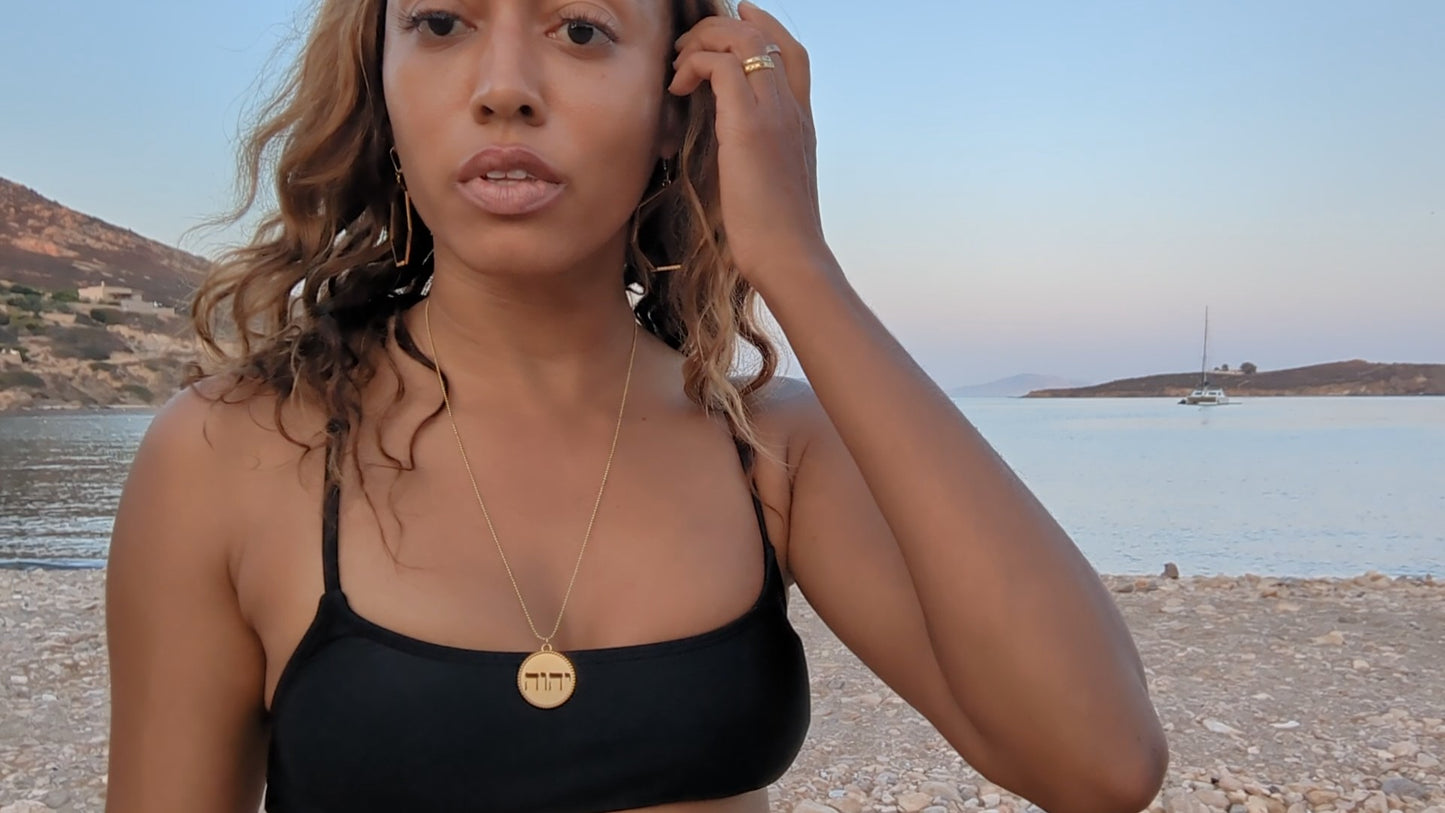 black woman on Greek beach in a black bikini wearing the gold Jehovah cutout coin from The Wandering Jewel