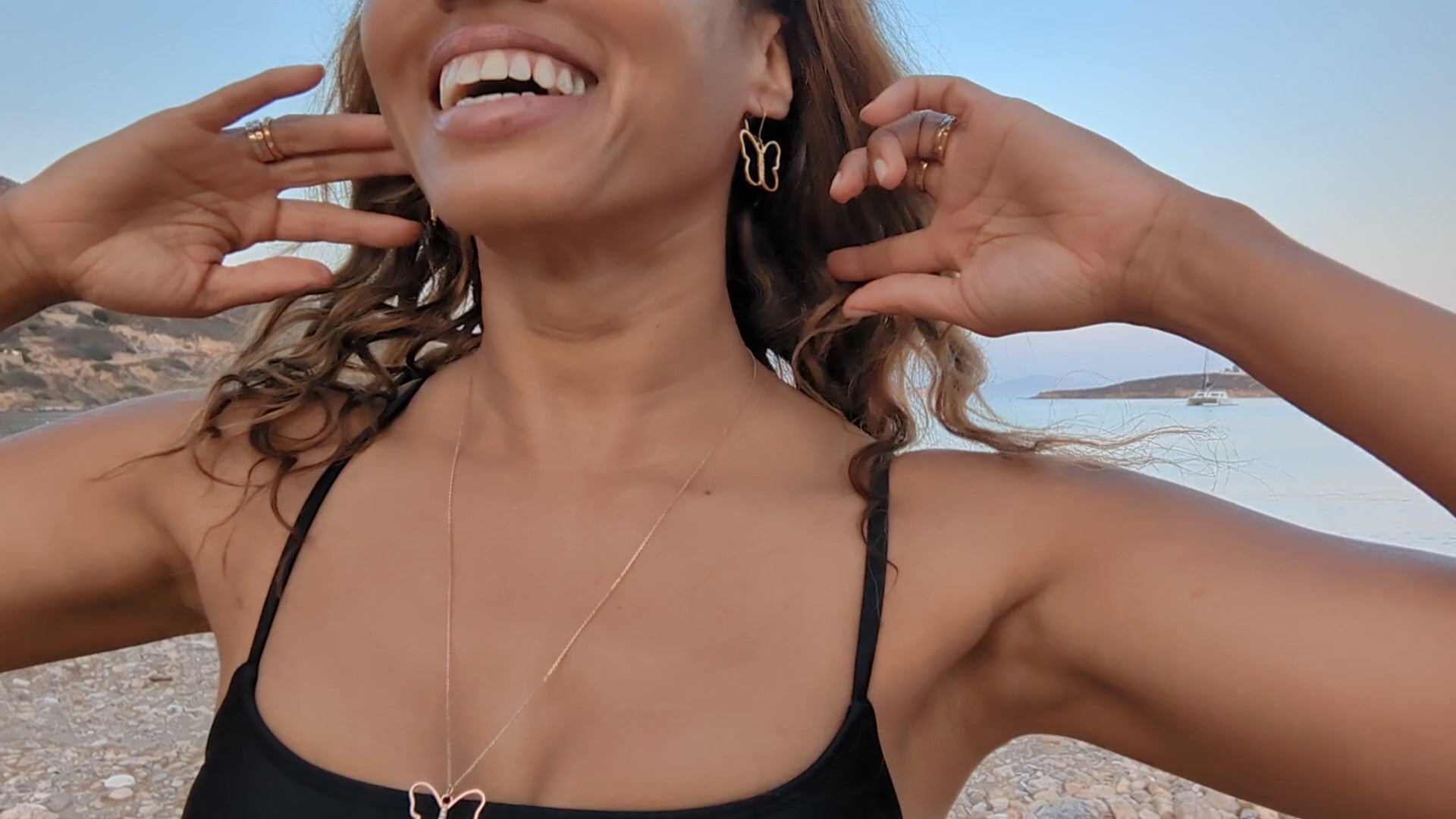 black woman in black bikini with her hands in the air on Greek beach wearing the 7 diamond butterfly earrings from the wandering jewel