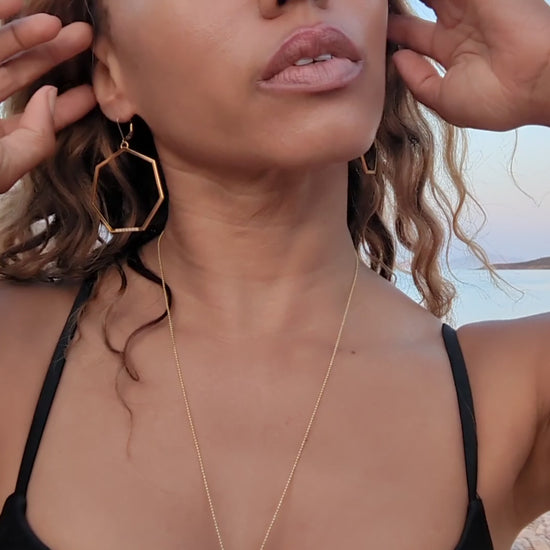 black woman in beach in a black bikini wearing Large gold hoop diamond earrings septagon shaped from the wandering jewel
