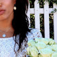 woman in bridal dress standing in front of white picket fence wearing flower pendant and matching earrings from the wandering jewel