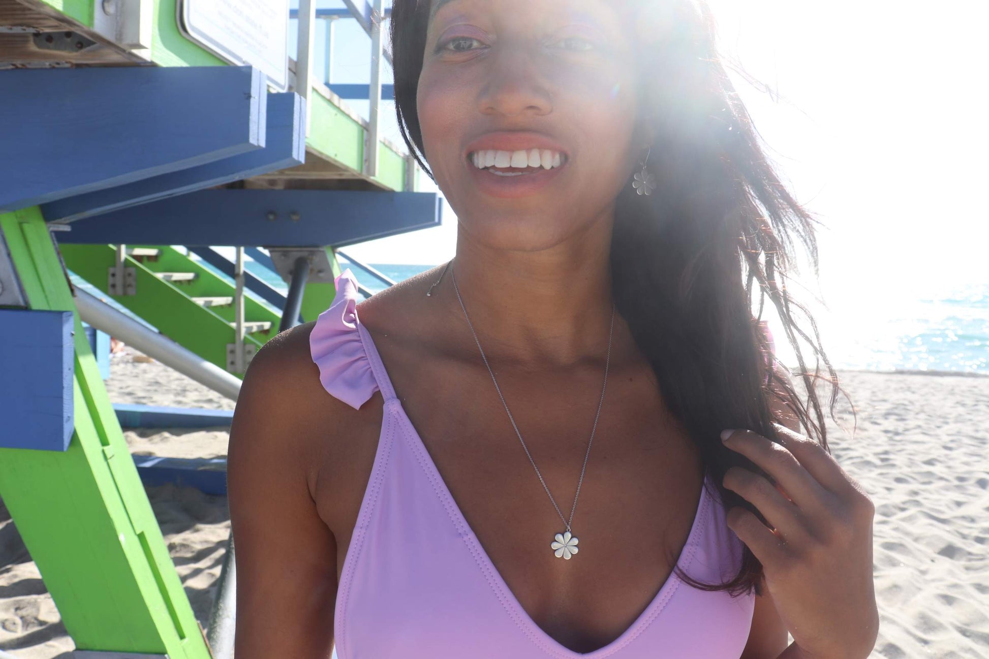 woman smiling on beach in a purple bikini wearing flower earrings and matching pendant