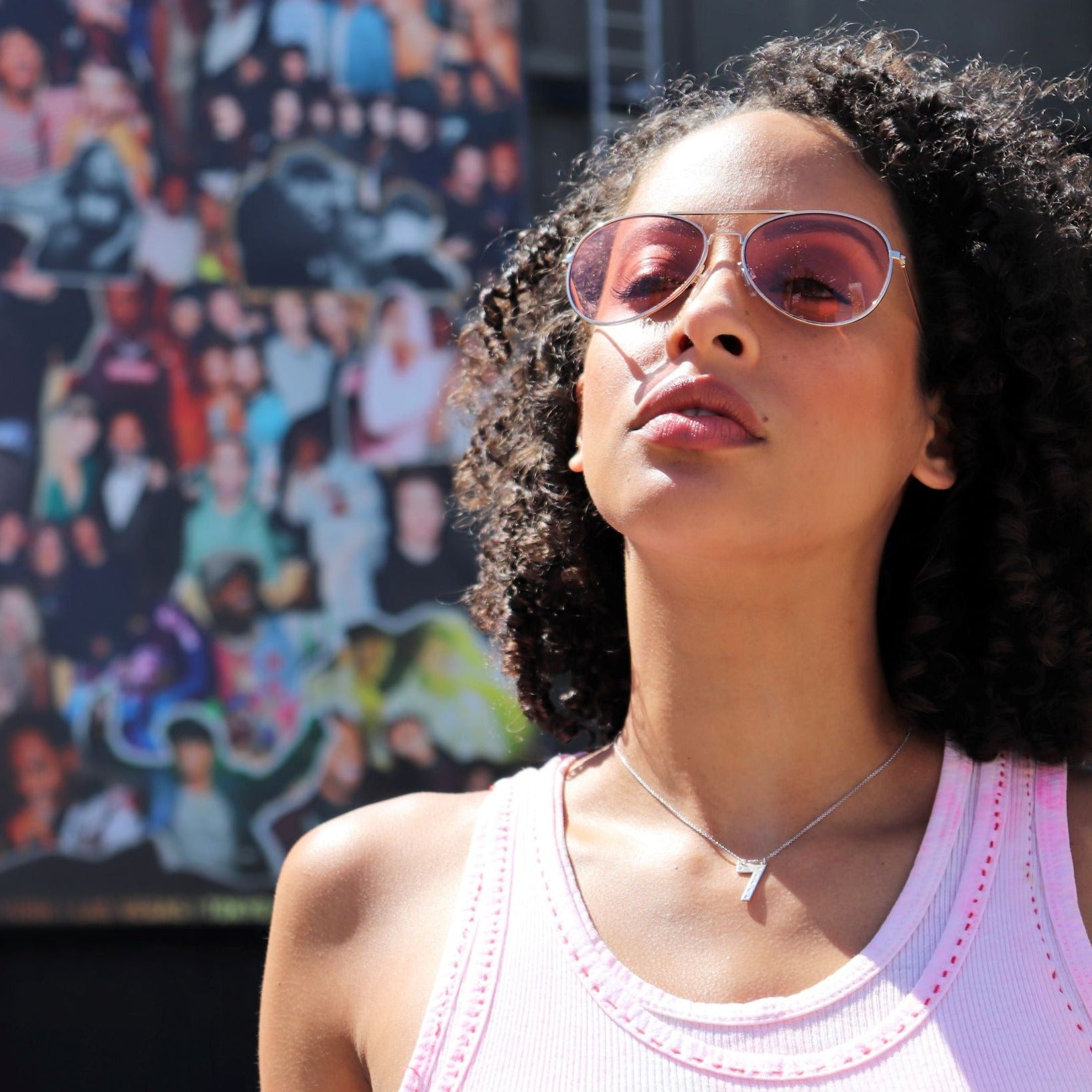 woman in pink tank top and pink sunglasses in front of a billboard wearing diamond number seven necklace