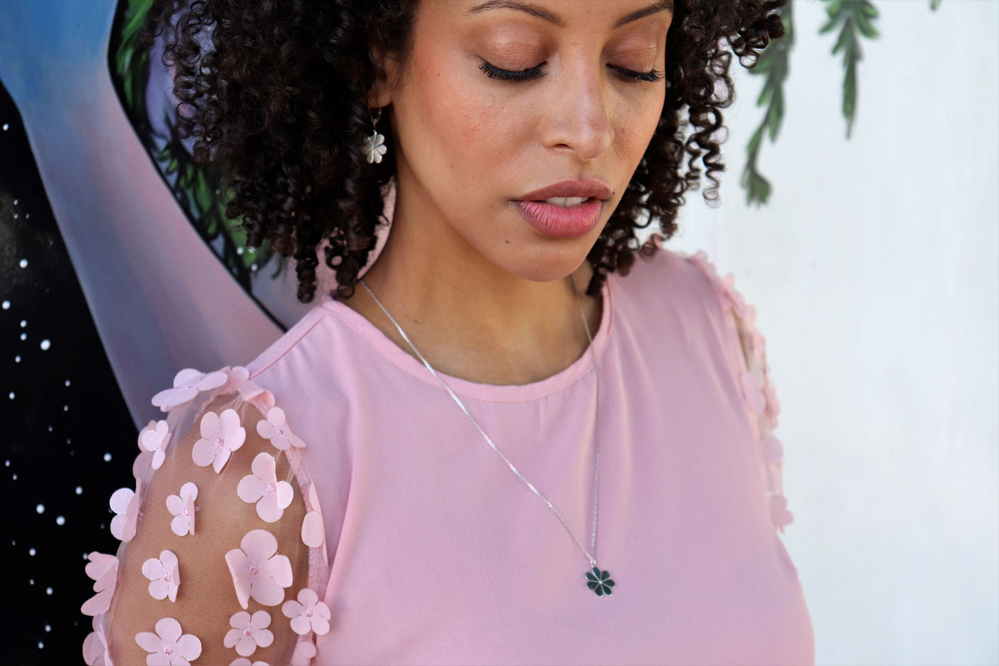 woman in pink dress standing in front of  graffiti mural wearing matching flower Earrings and necklace from the wandering jewel