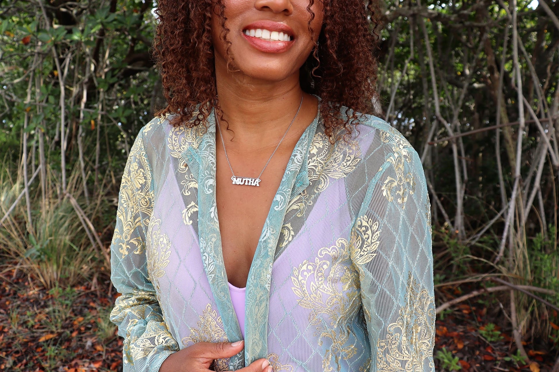 black woman in blue robe and purple tank top smiling in forest wearing mutha necklace from the wandering jewel