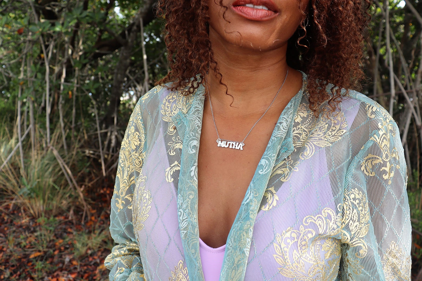 woman in forest wearing seafoam robe with a purple tank top wearing a nameplate mutha mother necklace