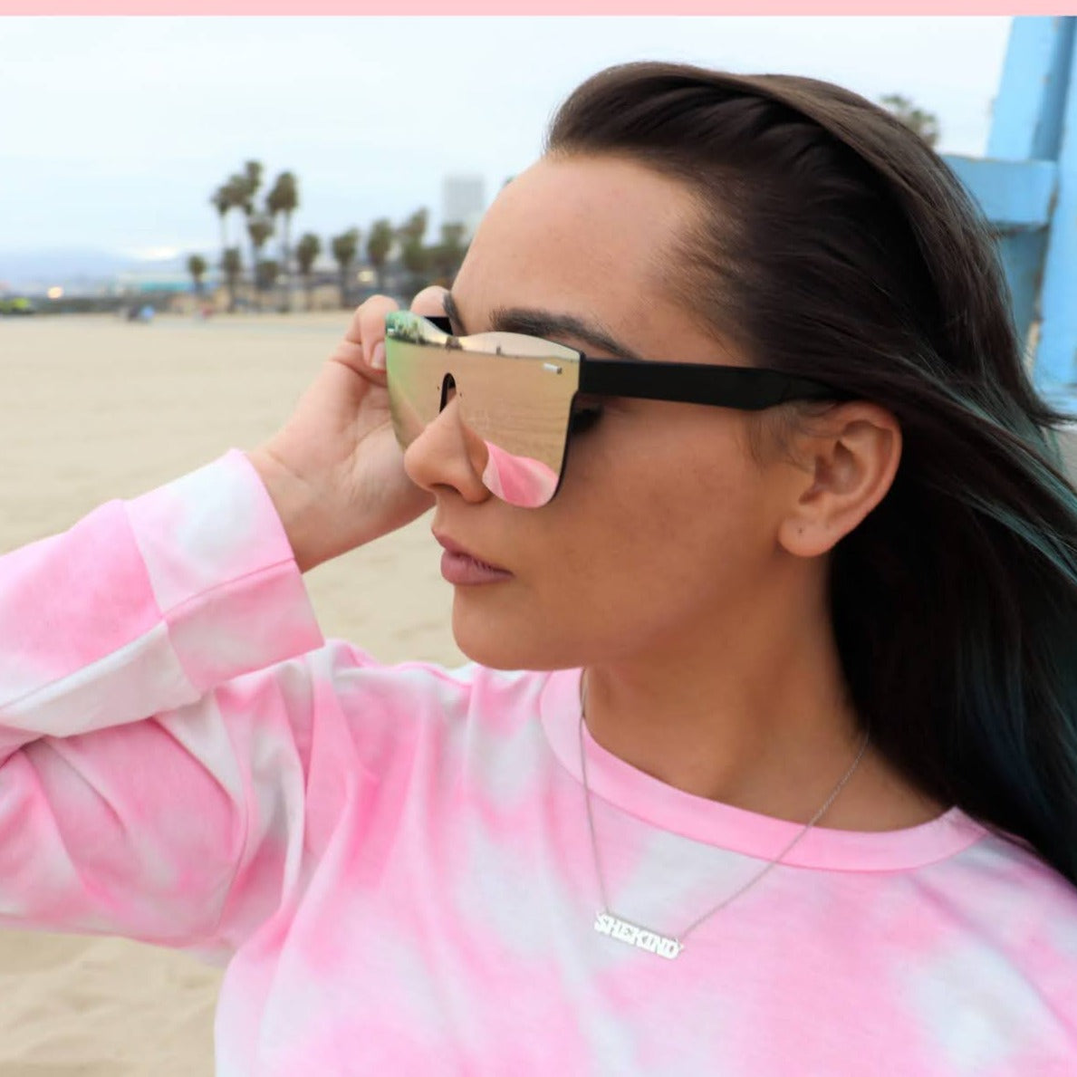 brunette woman on beach in pink tie dye shirt and sunglasses wearing a nameplate necklace that says shekind
