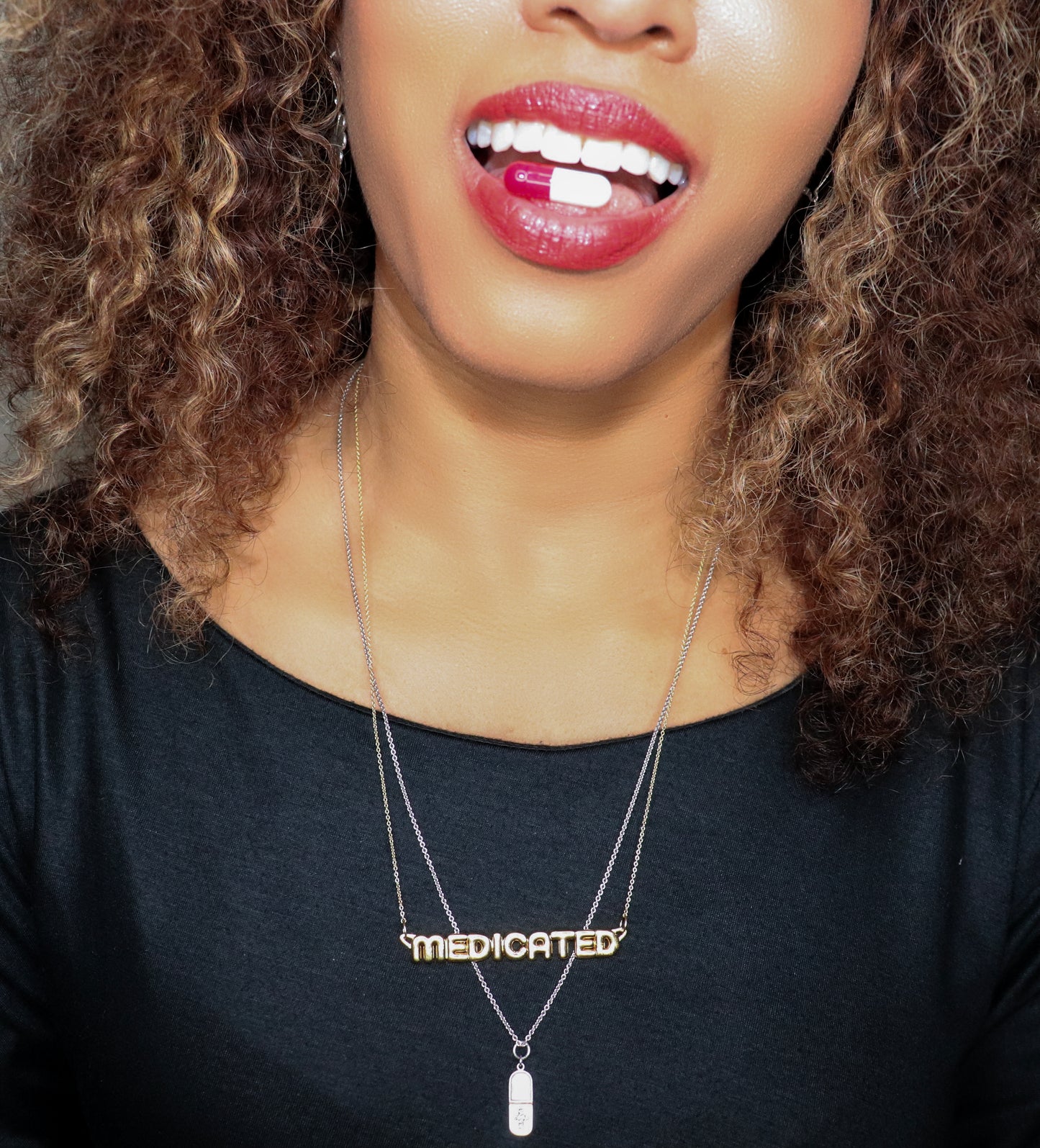 black woman with curly hair in a black dress with pill in her mouth wearing a necklace that says medicated