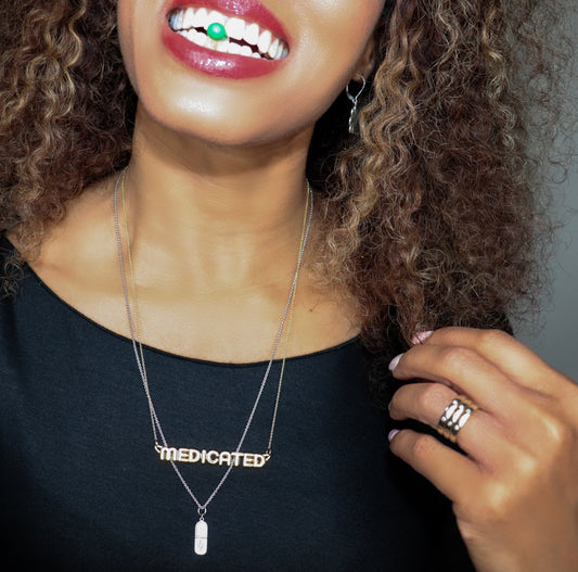 black woman in a black dress with green  pill in her mouth wearing a gold necklace that says medicated from the wandering jewel