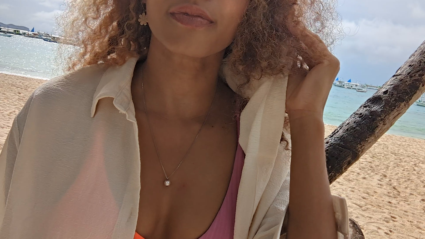 black woman in pink and orange bikini with white open blouse on beach wearing a white South Sea pearl pendant necklace from the wandering jewel