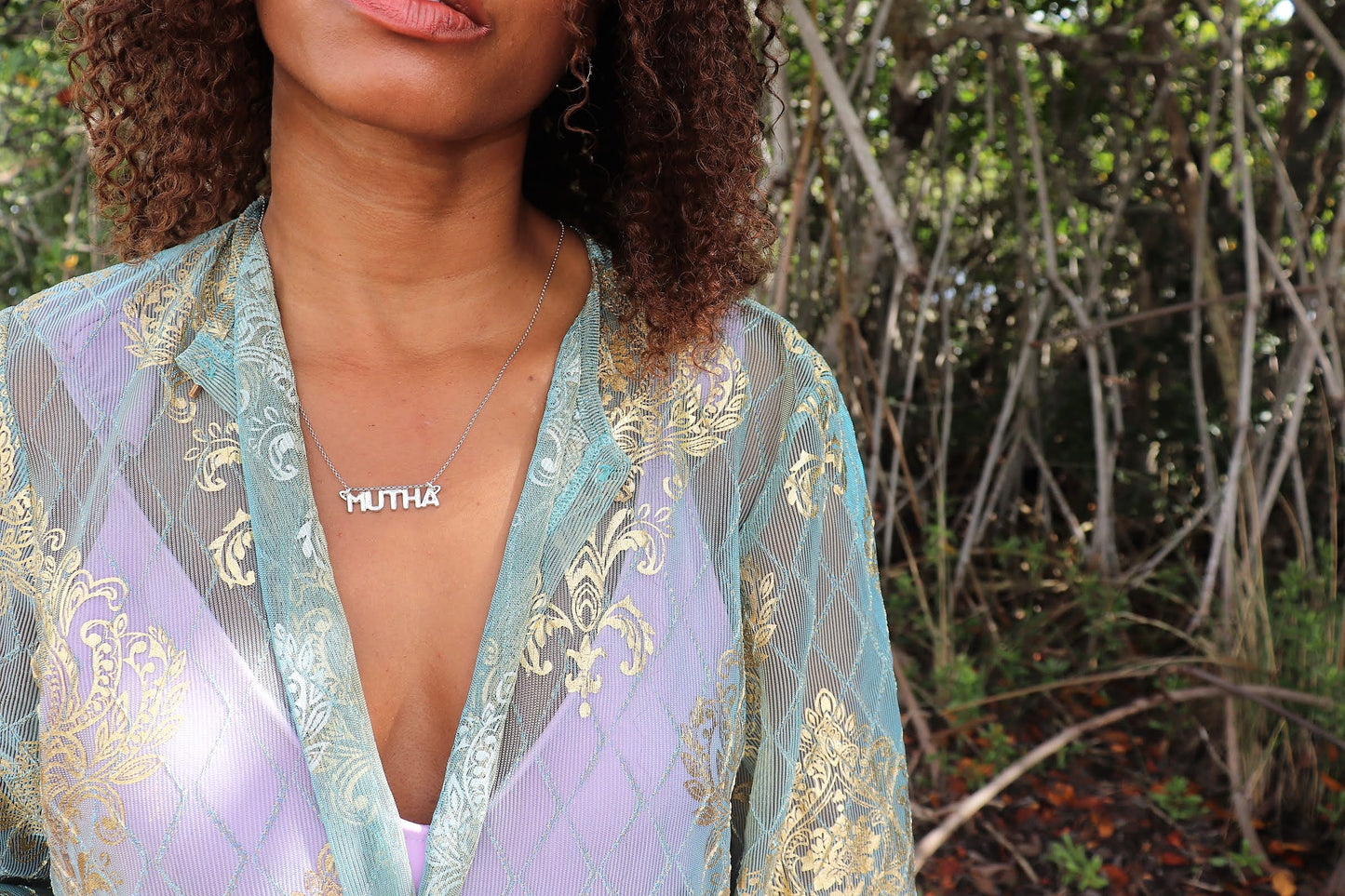 black woman in forest wearing a green and purple blouse over a lavender tank wearing mutha mother necklace