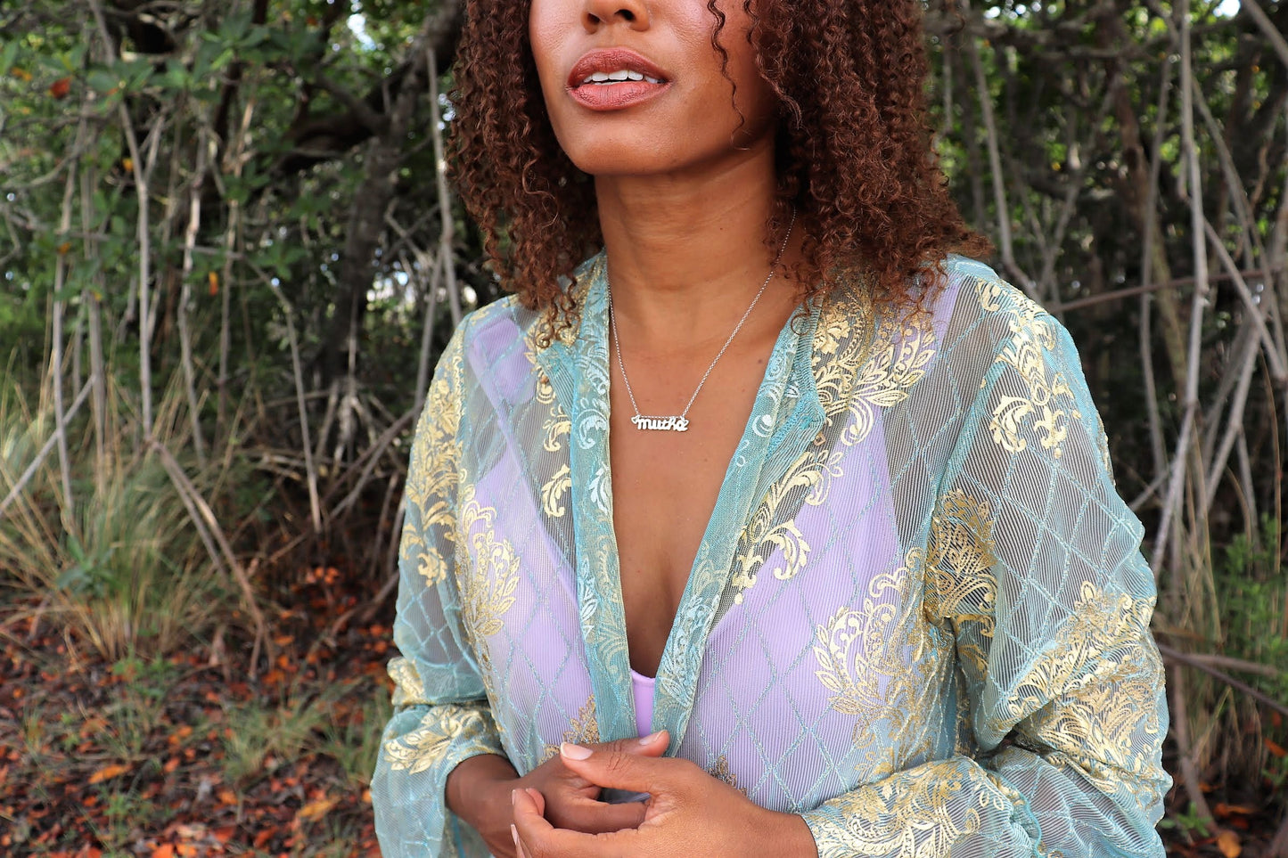 woman in forest smiling and wearing green and purple robe with a purple tank top and a  mutha mother necklace