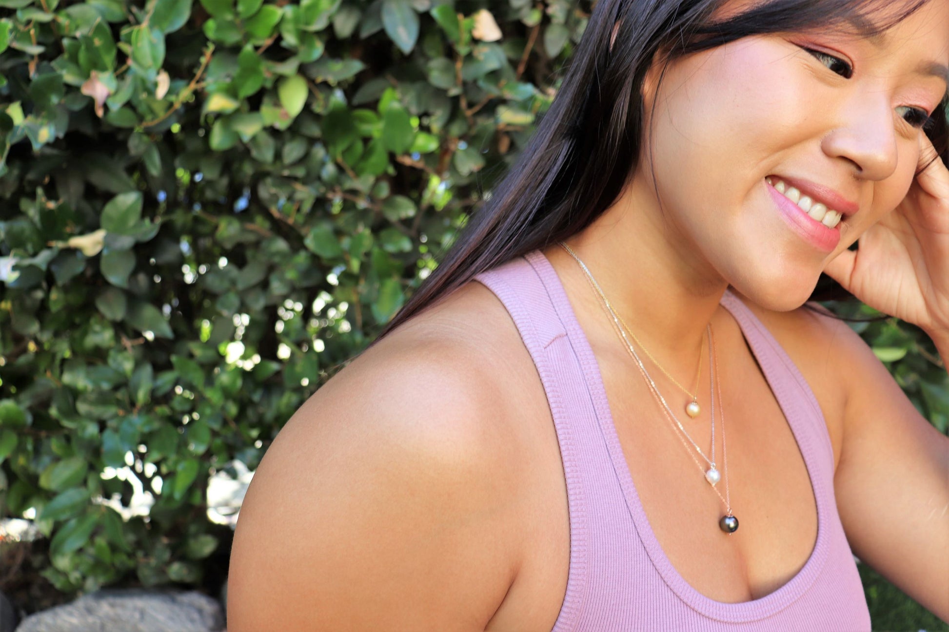 asian woman in purple tank top sitting in garden smiling and wearing three pearl pendant necklaces from the wandering jewel
