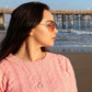 pretty brunette woman on beach in pink sweater in sunglasses looking to her left and wearing a  jade ring necklace from the wandering jewel