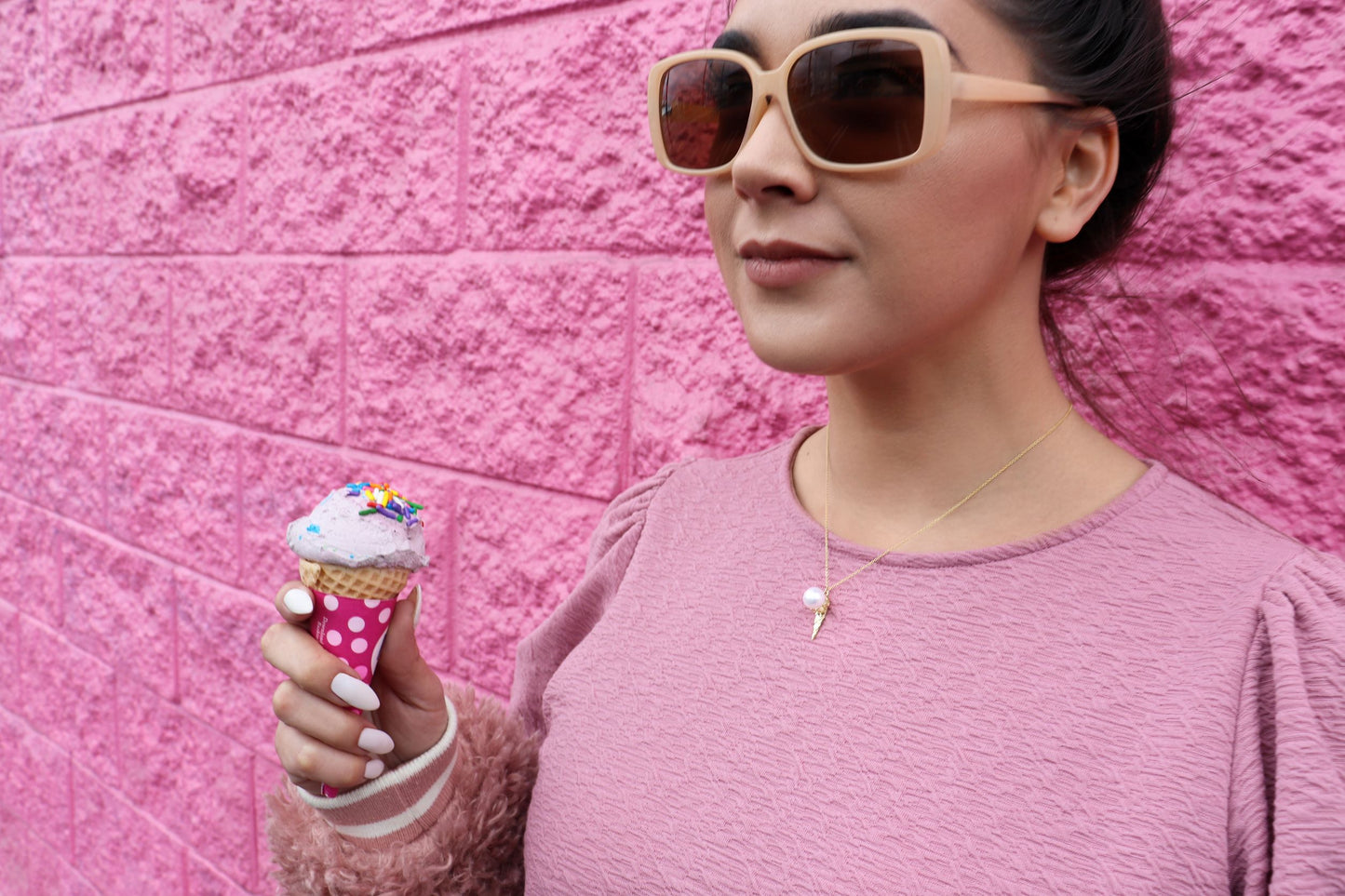 woman standing against a pink wall wearing a pink t-shirt and sunglasses eating ice cream and wearing a pearl ice cream pendant necklace