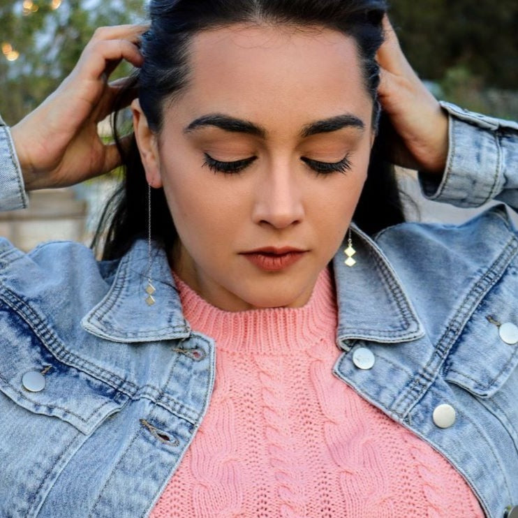 brunette woman with hands in her hair looking down wearing a pink shirt and denim jacket wearing dice earrings