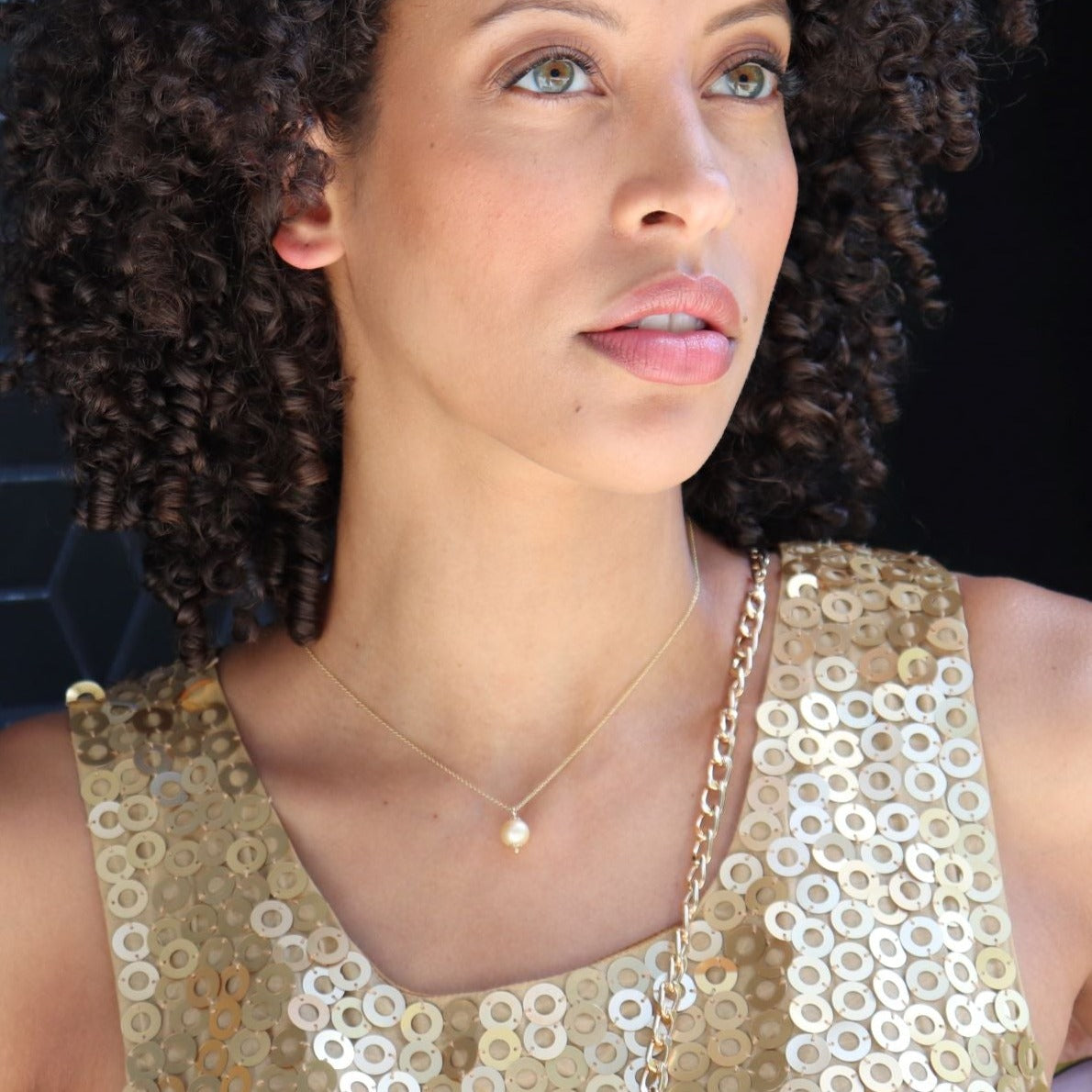 black woman with curly hair in gold sequin dress wearing gold pearl pendant