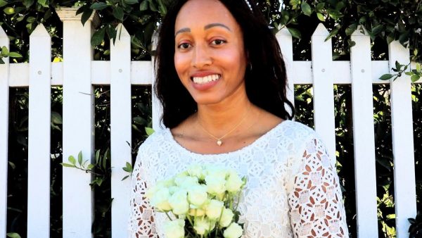 black woman in bridal dress standing in garden in front  of a white Pickett fence and wearing a the golden South sea pearl pendant from the wandering jewel