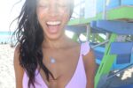 woman on beach in purple bikini smiling huge with her eyes closed wearing the yahweh coin pendant from The Wandering Jewel