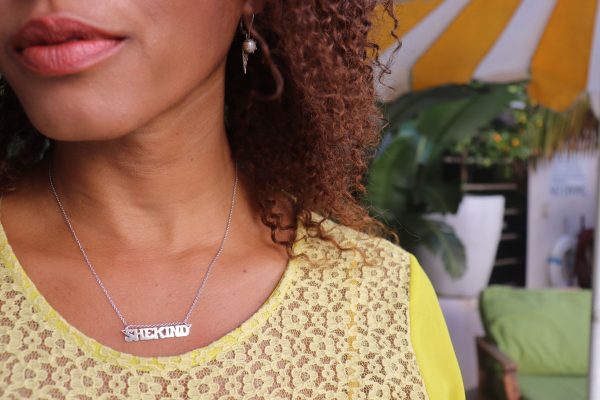 black woman with curly hair in yellow dress at pool wearing  a nameplate necklace that says shekind