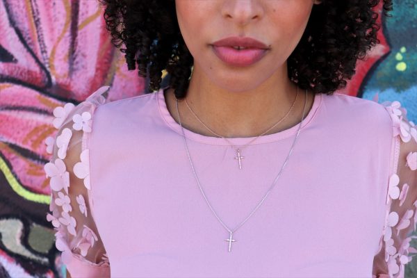 Woman wearing two crosses one is silver and the other is rose gold, both from the wandering jewel she is standing in front of a pink flower graffiti mural