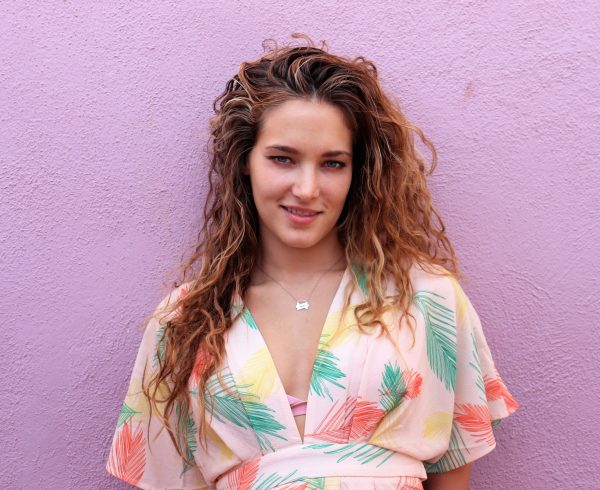 Blonde woman smiling and  wearing a multi color pink dress in front of pink building wearing a Double seven necklace from the wandering jewel