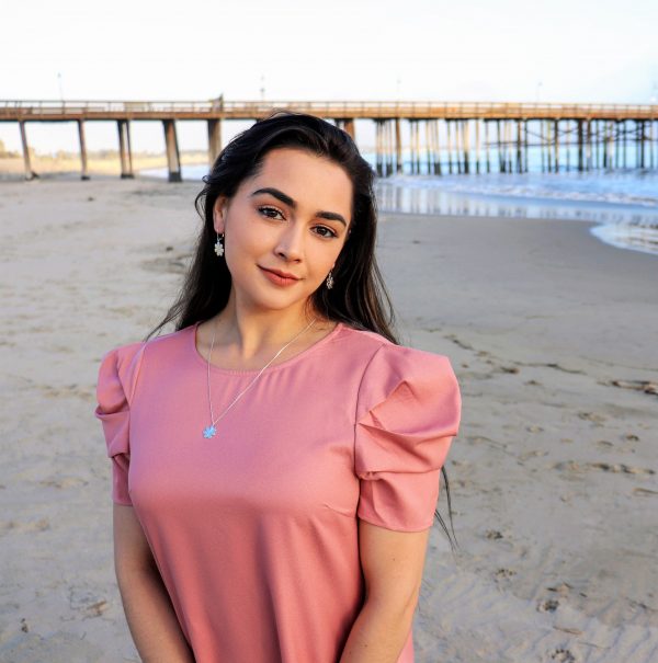 woman in a pink dress on beach wearing flower earrings and a matching necklace from the wandering jewel