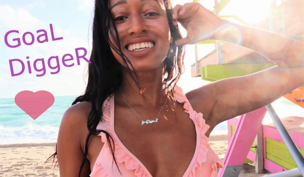 black woman on beach in pink bikini in front of a lifeguard stand wearing goaldigger necklace