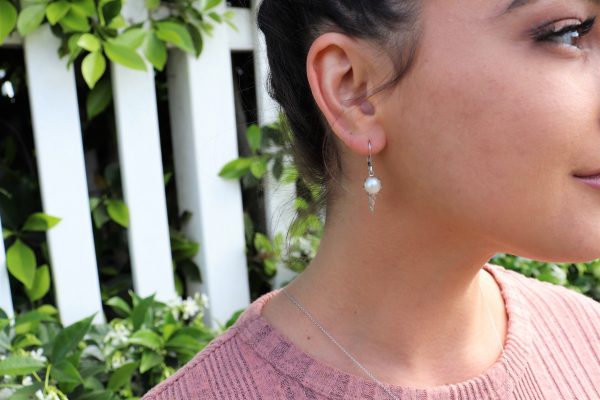 brunette woman in pink top in front of a white picket fence in a garden wearing ice cream pearl pendant earrings from the wandering jewel