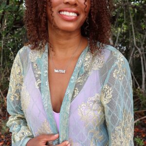 black woman in blue robe and purple tank top smiling in forest wearing mutha necklace from the wandering jewel