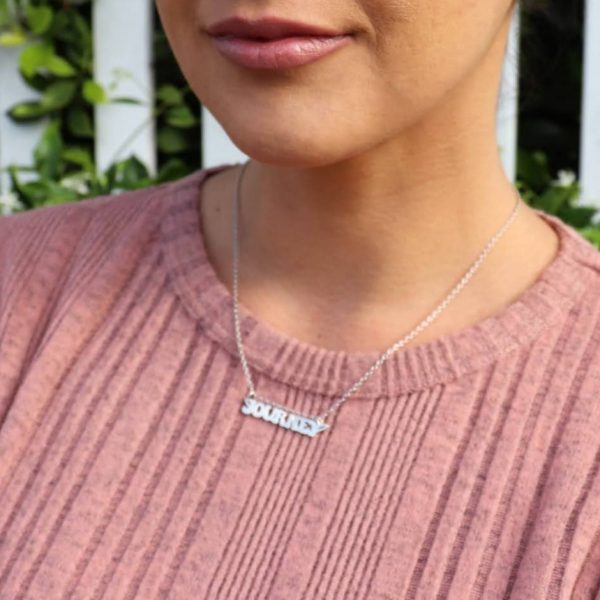 woman in pink shirt standing in front of a white picket fence in a garden wearing nameplate journey necklace