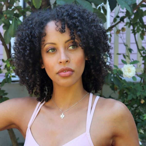 Woman in white sports bra sitting in front of garden wearing diamond love knot necklace