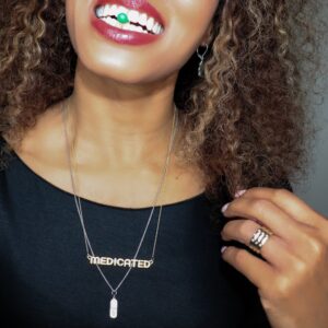 black woman in a black dress with green  pill in her mouth wearing a gold necklace that says medicated from the wandering jewel