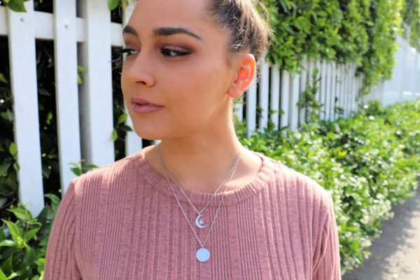 Woman with pink top standing in front of a white fence and garden wearing a silver moon and star coin necklace from the wandering jewel