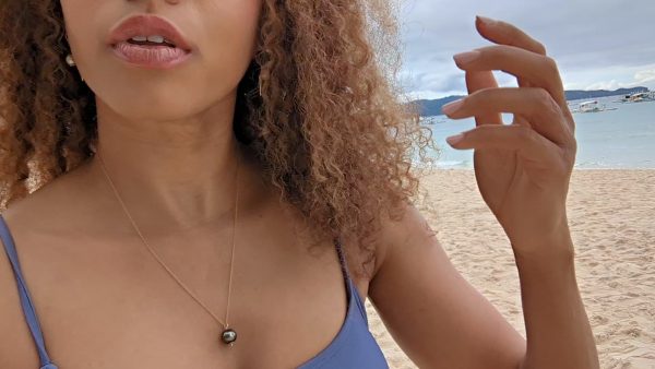 black woman with curly hair in a purple bikini on a beach wearing the black South sea pearl pendant from the wandering jewel