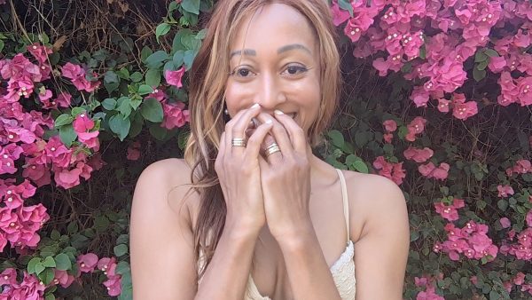 woman smiling in light brown tank top and standing in front of bright pink flowers wearing 6  18K solid gold disengagement rings from the wandering jewel