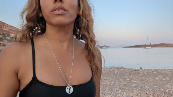 black woman in black bikini on Greek beach wearing  the cross cutout coin from the wandering jewel