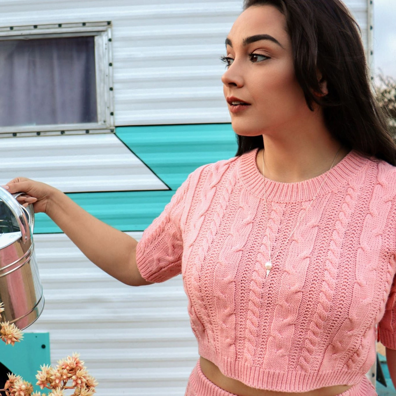 woman in pink sweater and skirt on front of blue and white trailer watering plants and wearing pearl ice cream cone pendant necklace from the wandering jewel