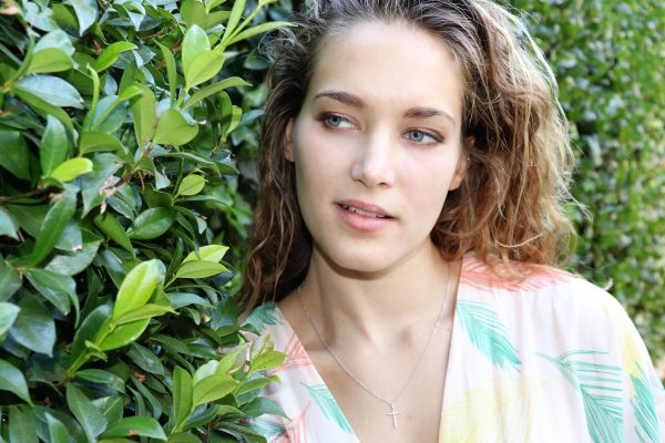 Woman in pink multi color dress in a garden wearing small silver cross necklace  from the wandering jewel